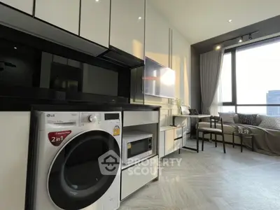 Modern living room with integrated kitchen and washer, featuring sleek cabinetry and large window.