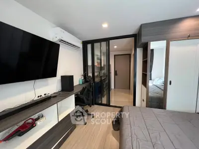 Modern bedroom with sleek design, featuring a large TV, workspace, and sliding glass doors leading to a cozy kitchen.