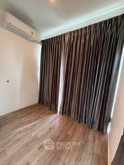Spacious room with wooden flooring and large window with dark curtains, featuring air conditioning.
