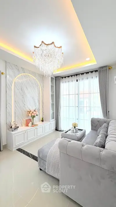Luxurious living room with elegant chandelier and plush gray sofa, featuring marble accent wall and large windows.