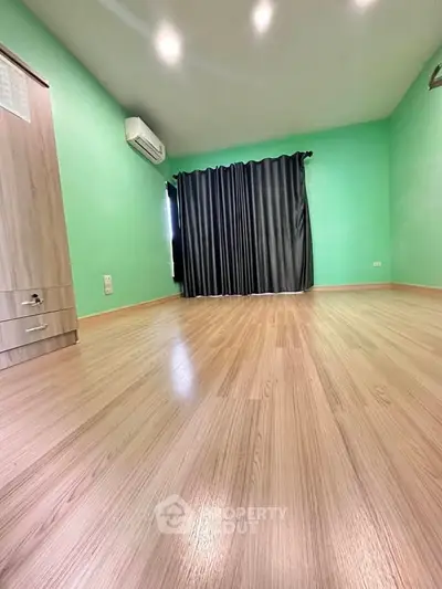 Spacious empty room with wooden flooring and green walls, featuring air conditioning and dark curtains.