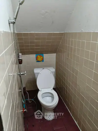 Compact bathroom with sloped ceiling and tiled walls, featuring a toilet and handheld shower.