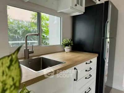 Modern kitchen with sleek black fridge and wooden countertop, featuring a stainless steel sink and lush greenery view.