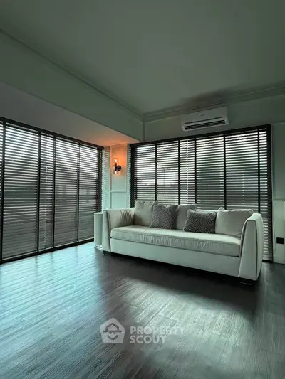 Spacious living room with modern sofa and large windows with blinds, featuring sleek wooden flooring.