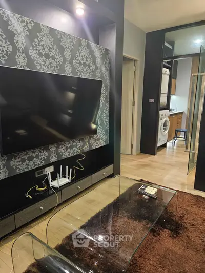 Modern living room with stylish wallpaper and large TV, leading to a kitchen with laundry appliances.