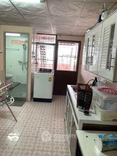 Compact kitchen with washing machine and bathroom access, featuring tiled flooring and ample storage cabinets.