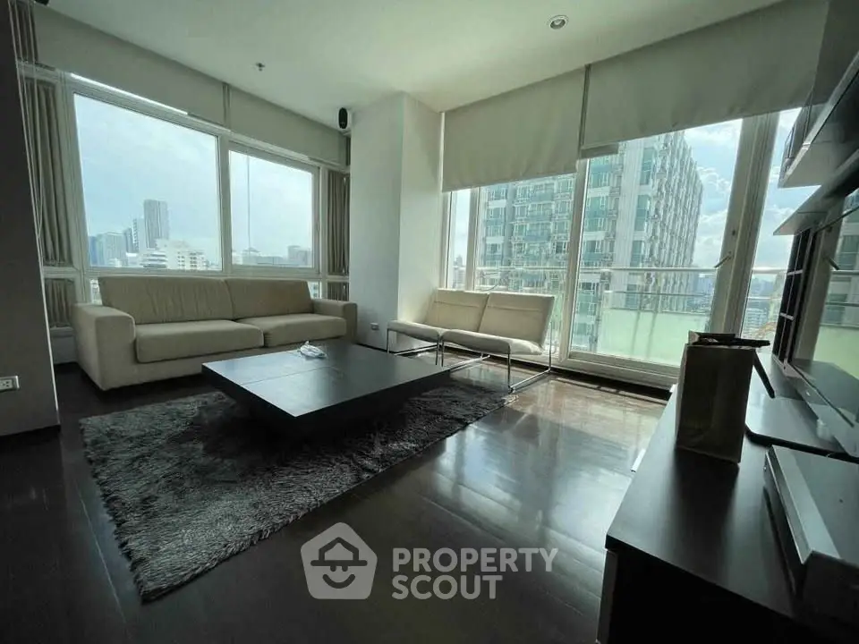 Spacious modern living room with large windows and city view, featuring sleek furniture and dark wood flooring.