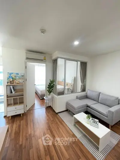 Modern living room with gray sofa, wooden flooring, and view into a cozy bedroom.