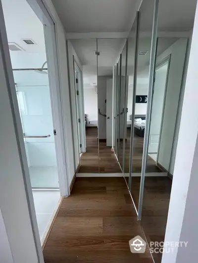 Modern hallway with mirrored closets and wooden flooring leading to a bright bedroom.