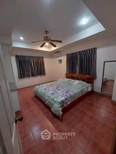 Spacious bedroom with terracotta tiled flooring, a large bed with patterned bedding, ceiling fan, and ample natural light from curtained windows.