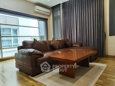 Modern living room with leather sofa and wooden coffee table, featuring large windows and elegant curtains.