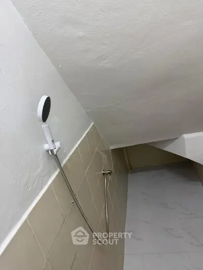 Compact bathroom with sloped ceiling and handheld showerhead, featuring beige tiles and white flooring.