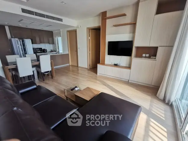 Spacious modern living room with open kitchen and natural light, featuring sleek furniture and wooden flooring.