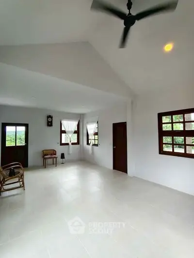 Spacious living room with high ceiling and natural light, featuring modern fan and large windows.