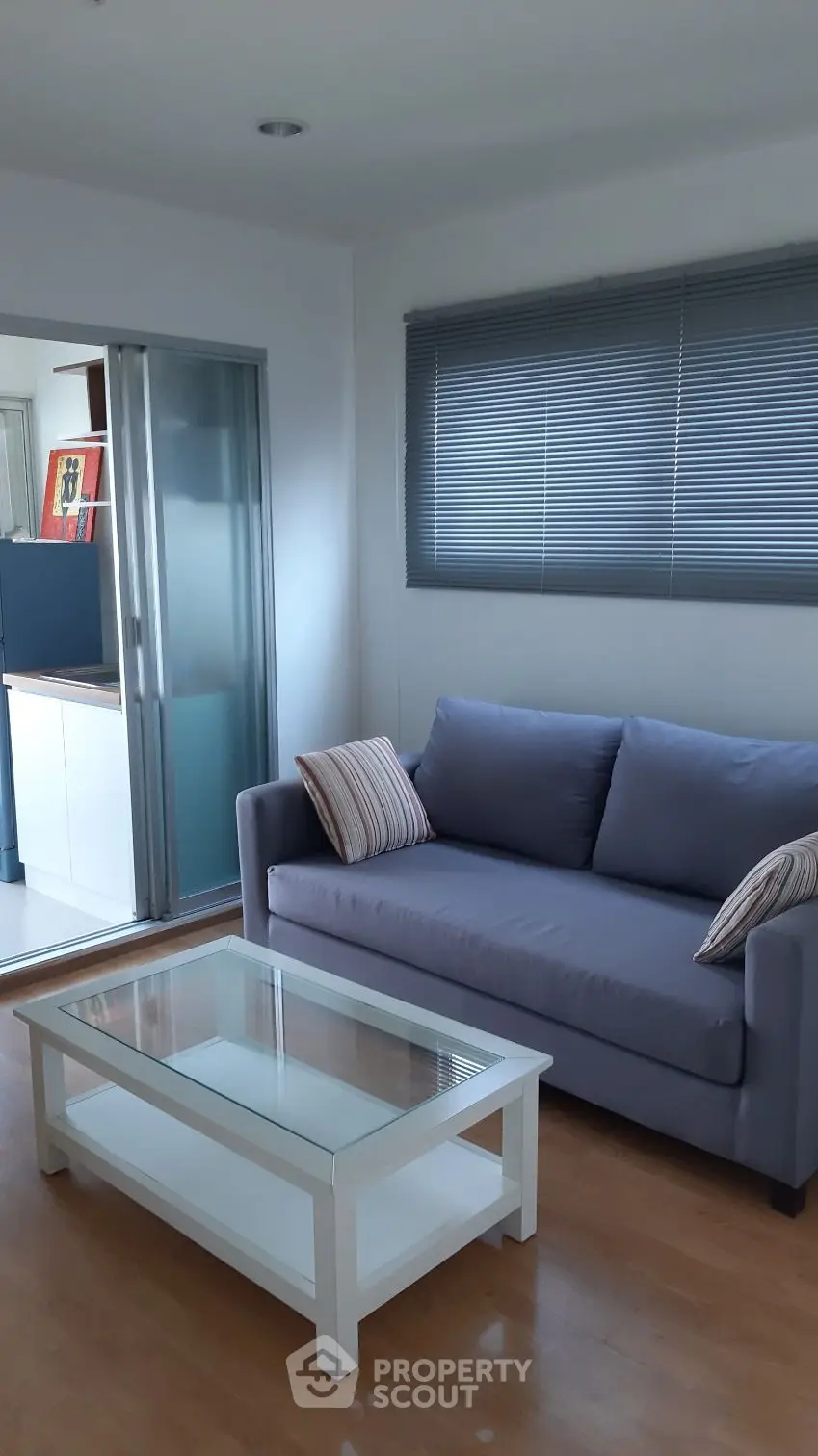 Modern living room with sleek sofa and glass coffee table, featuring a sliding door to a bright kitchen area.