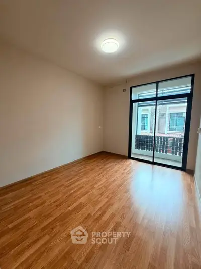 Spacious empty room with wooden flooring and large window leading to balcony.