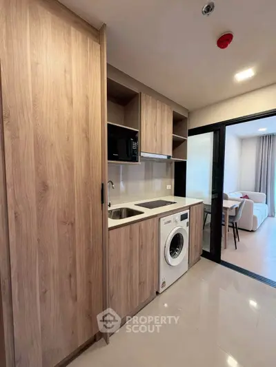 Modern kitchen with wooden cabinets, washing machine, and open layout leading to living area.