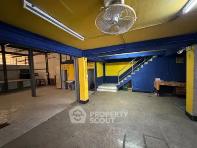Spacious industrial-style interior with vibrant yellow and blue walls, featuring a staircase and ceiling fan.