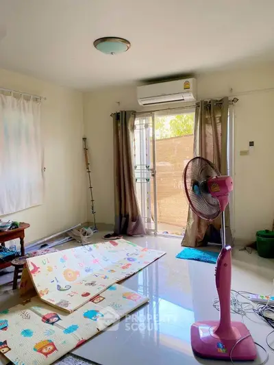 Bright and airy living space with ample natural light, featuring a large window with curtains, an air conditioning unit, and a playful children's play mat.