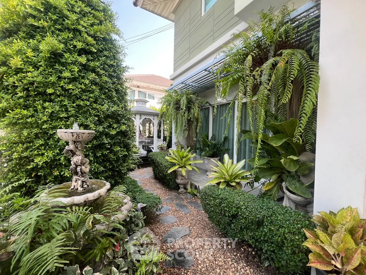 Charming garden path with lush greenery and elegant fountain beside modern home exterior.