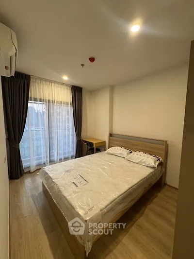 Cozy bedroom with wooden flooring and large window, featuring a comfortable bed and study desk.