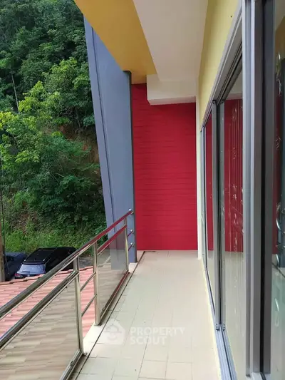 Spacious balcony with glass railing and vibrant red accent wall, offering serene green views.