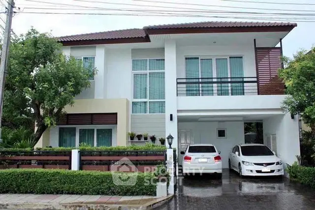 Modern two-story house with two cars parked in driveway, lush greenery, and large windows.
