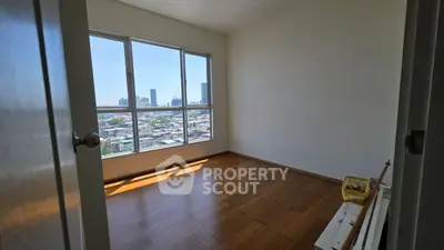 Bright empty room with wooden floors and large window offering city skyline view.