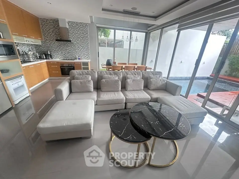 Luxurious open-plan living room with modern kitchen and pool view, featuring elegant furniture and large windows.