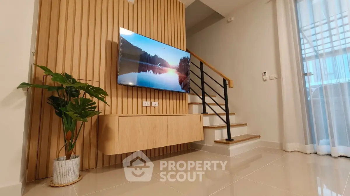 Modern living room with stylish wooden paneling and mounted TV, elegant staircase, and large window.