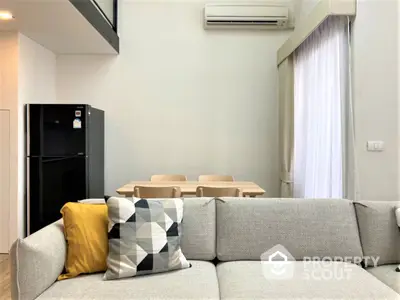 Modern minimalist living room with a comfortable grey sectional sofa, vibrant throw pillows, sleek black refrigerator, and a cozy dining area with wooden furniture.