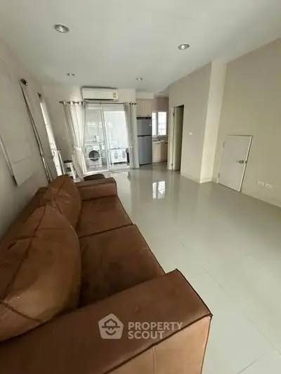 Spacious living room with modern kitchen and brown sofa, featuring sleek tile flooring and natural light.