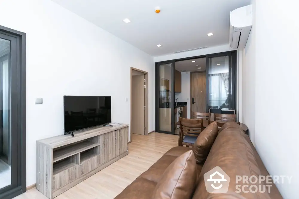 Modern living room with leather sofa and TV, open to sleek kitchen area.