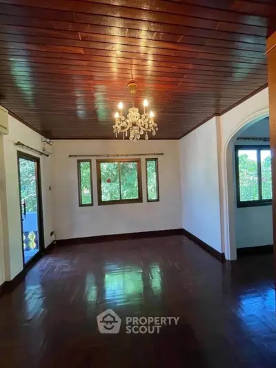 Charming living room with wooden ceiling and chandelier, featuring large windows with lush green views.