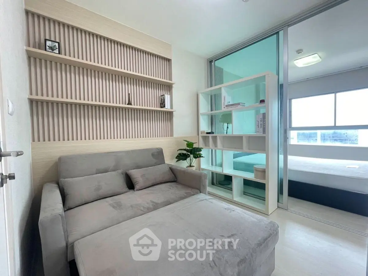 Modern living room with cozy gray sofa and stylish shelving, adjacent to a bright bedroom with sliding glass doors.