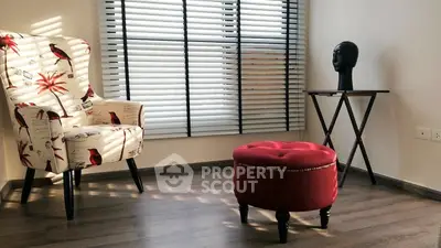 Chic modern living room with stylish armchair, red ottoman, and natural light through blinds.