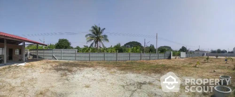 Expansive vacant land with potential for development, surrounded by a secure fence and lush greenery, under a clear blue sky.