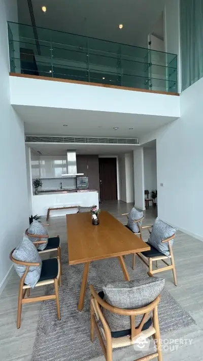 Spacious double-height living room with modern open kitchen, elegant dining set, and a cozy upper-level loft area with glass balustrade, offering a luxurious and airy ambiance.