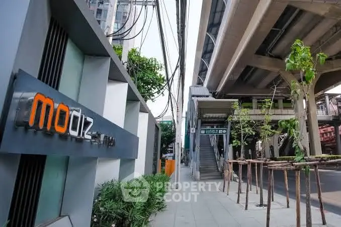 Modern urban building entrance near elevated train tracks and lush greenery.