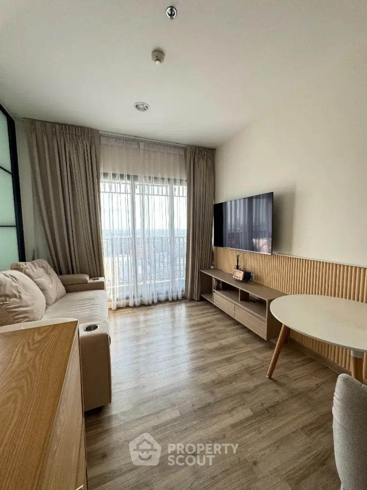 Modern living room with large TV and balcony view, featuring stylish furniture and wooden flooring.