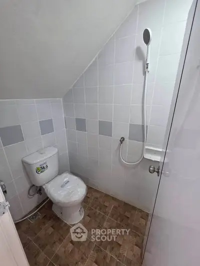 Compact bathroom with shower and toilet under sloped ceiling, featuring tiled walls and floor.