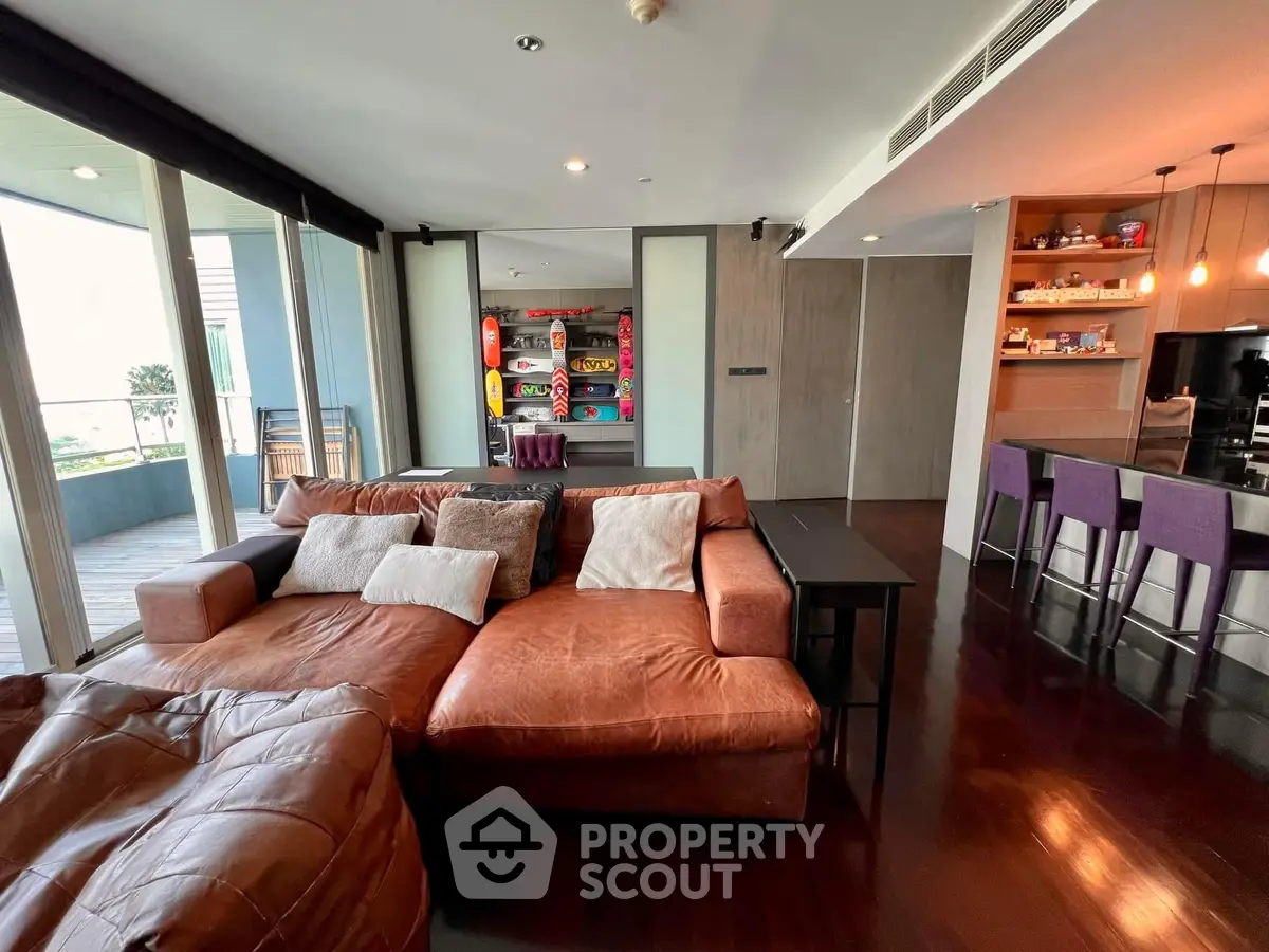 Spacious open-plan living area with plush leather sofa, sleek hardwood floors, and modern kitchen with bar seating, leading to a sunlit balcony.