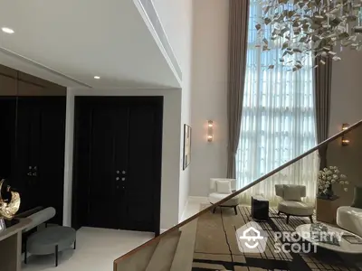 Opulent living room with high ceilings, elegant chandelier, and modern decor, featuring a grand staircase and floor-to-ceiling windows with sheer curtains.