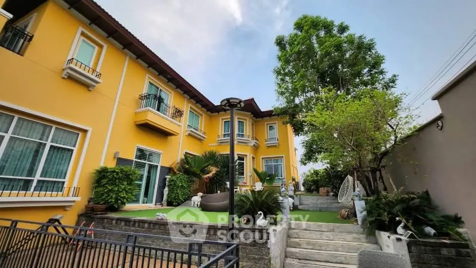 Charming yellow two-story house with lush garden and elegant outdoor seating area.