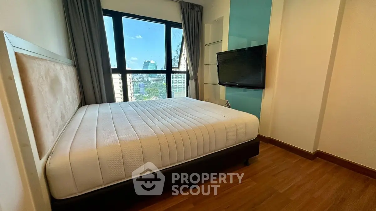 Modern bedroom with large window offering city view, featuring a comfortable bed and mounted TV.
