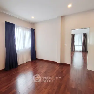 Spacious living room with polished hardwood floors, ample natural light, and elegant blue curtains, leading to an adjoining room through an open doorway.