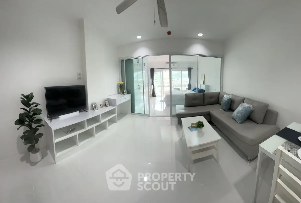 Modern living room with sleek white furniture and open layout, leading to a bright bedroom with a view.