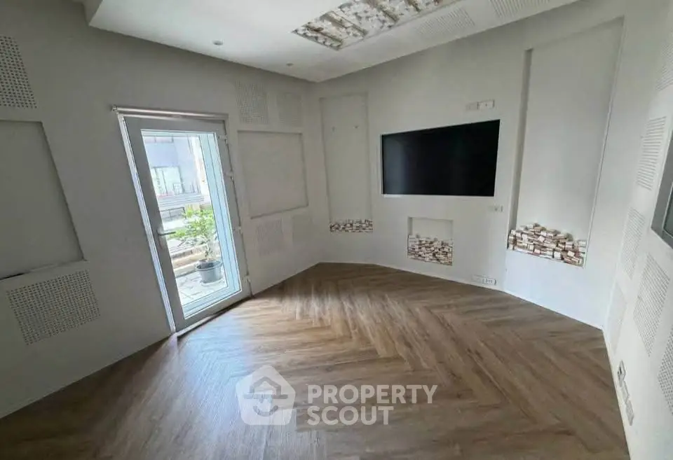 Modern living room with wooden flooring and large TV, leading to a balcony with outdoor view.