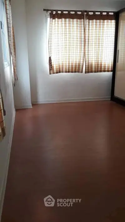 Spacious empty room with wooden flooring and large windows with curtains.