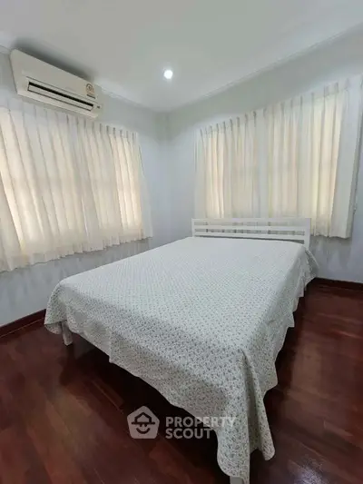 Cozy bedroom with wooden flooring and air conditioning, featuring soft natural light through curtains.